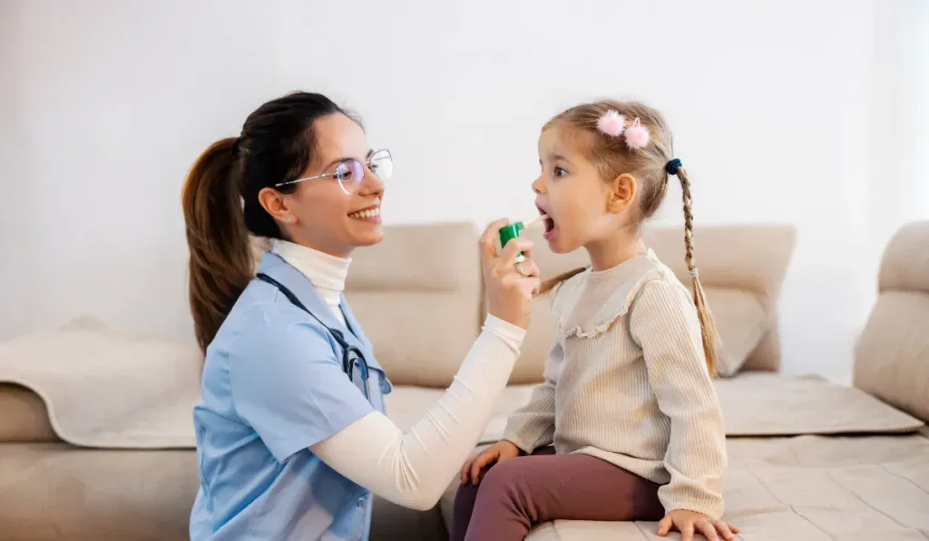 Caring female pediatrician spraying medicine into sick little girl's throat and helping her heal flu during her home visit.
